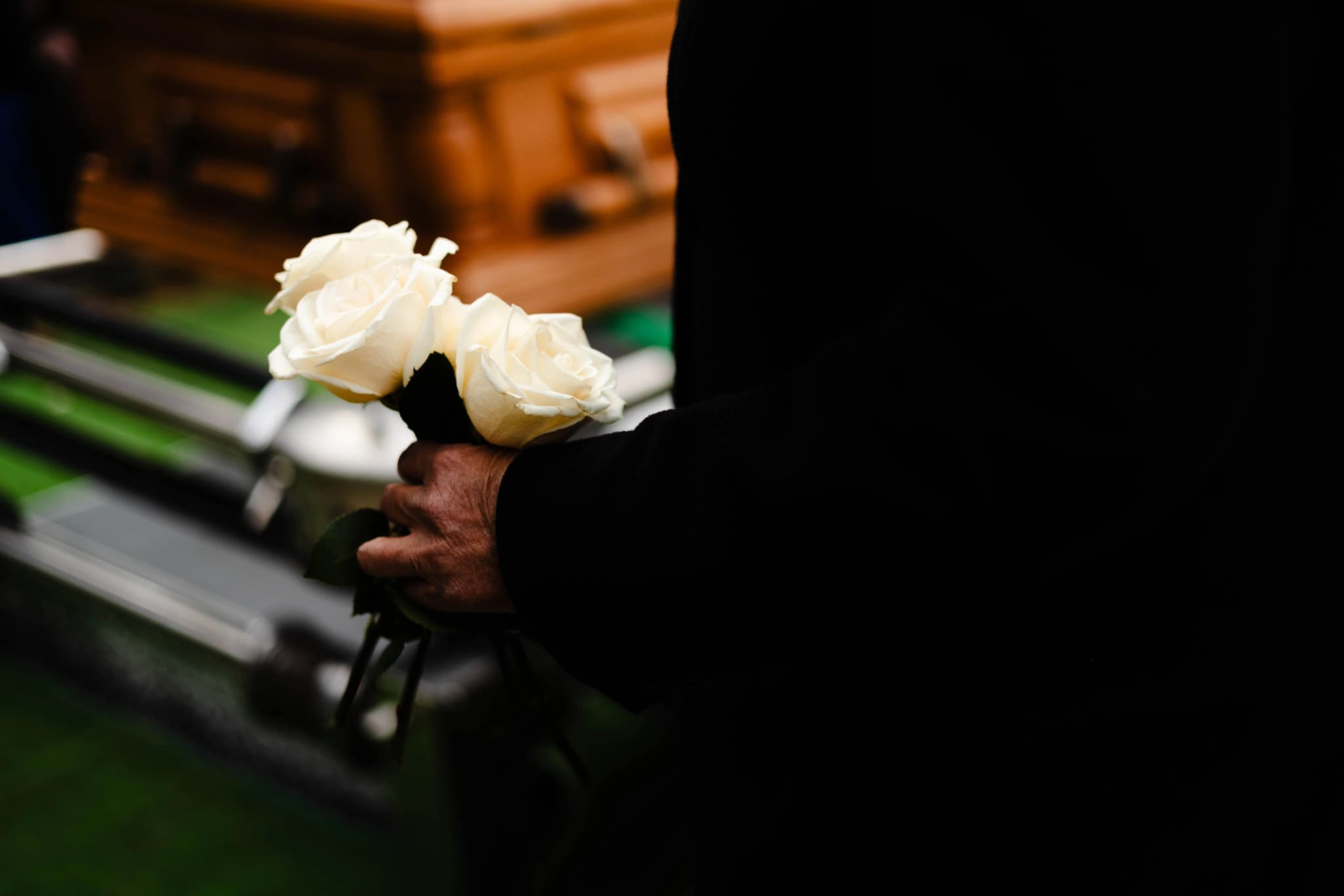 A hand in a black suit holding three white roses in front of a wooden casket at a funeral service.